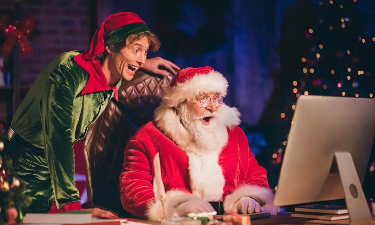 Санта и эльф с волнением проверяют компьютер, планируя торговые ходы и праздничные расписания на конец года.