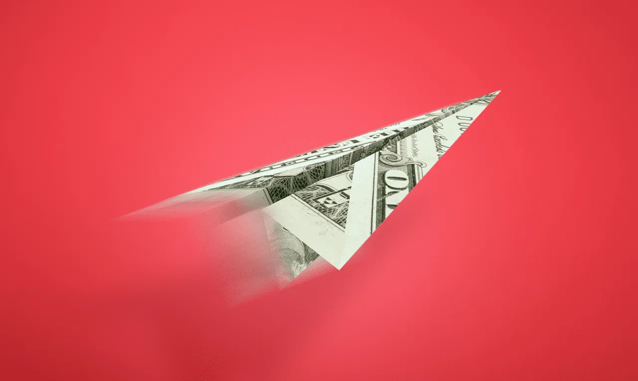 طائرة ورقية مصنوعة من ورقة الدولار تطير إلى الأمام على خلفية حمراء، مما يرمز إلى النمو المالي أو المخاطر.