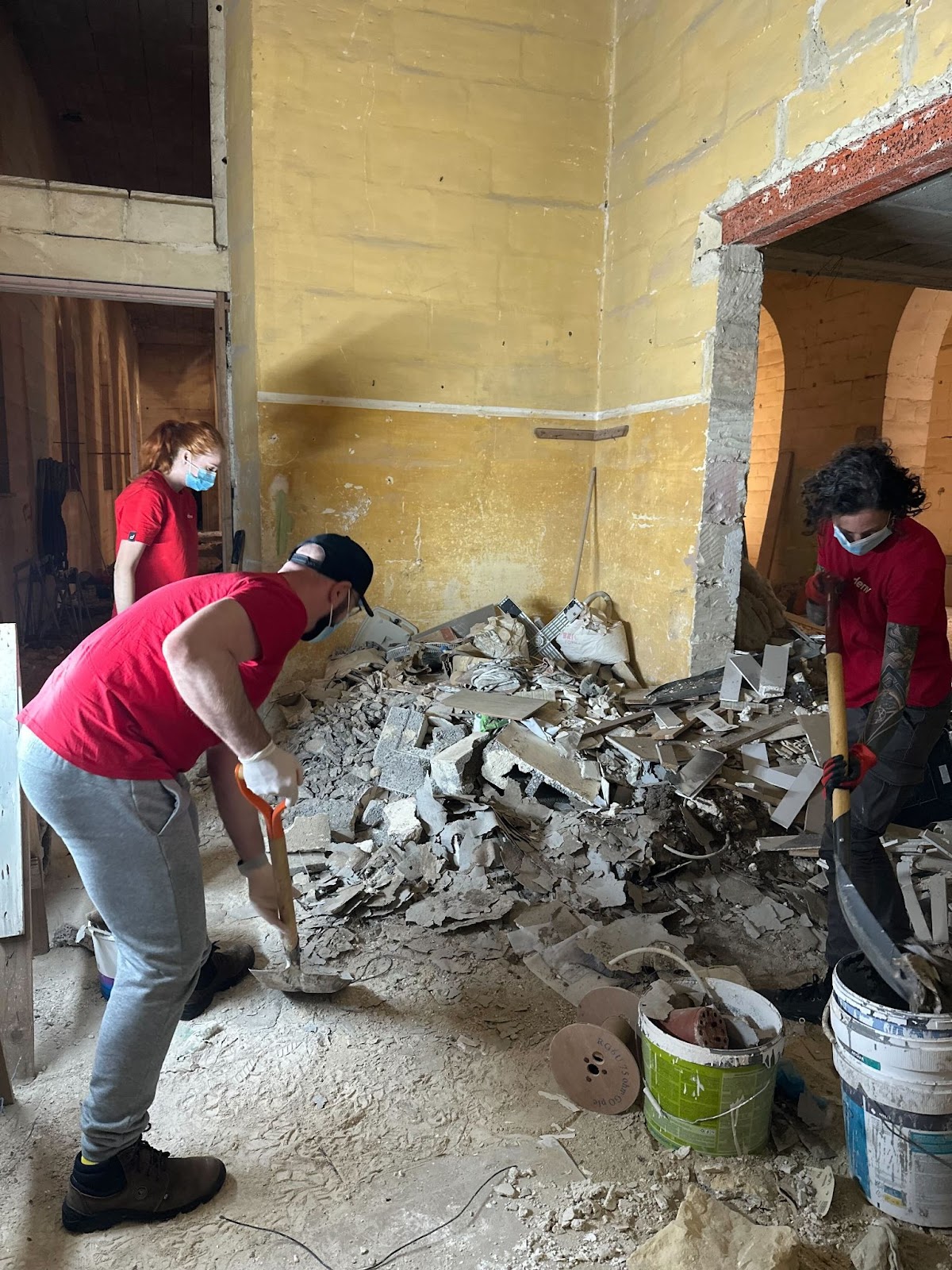 Deriv volunteers clear rubble and debris inside a damaged building as part of a renovation effort, wearing red shirts and face masks.